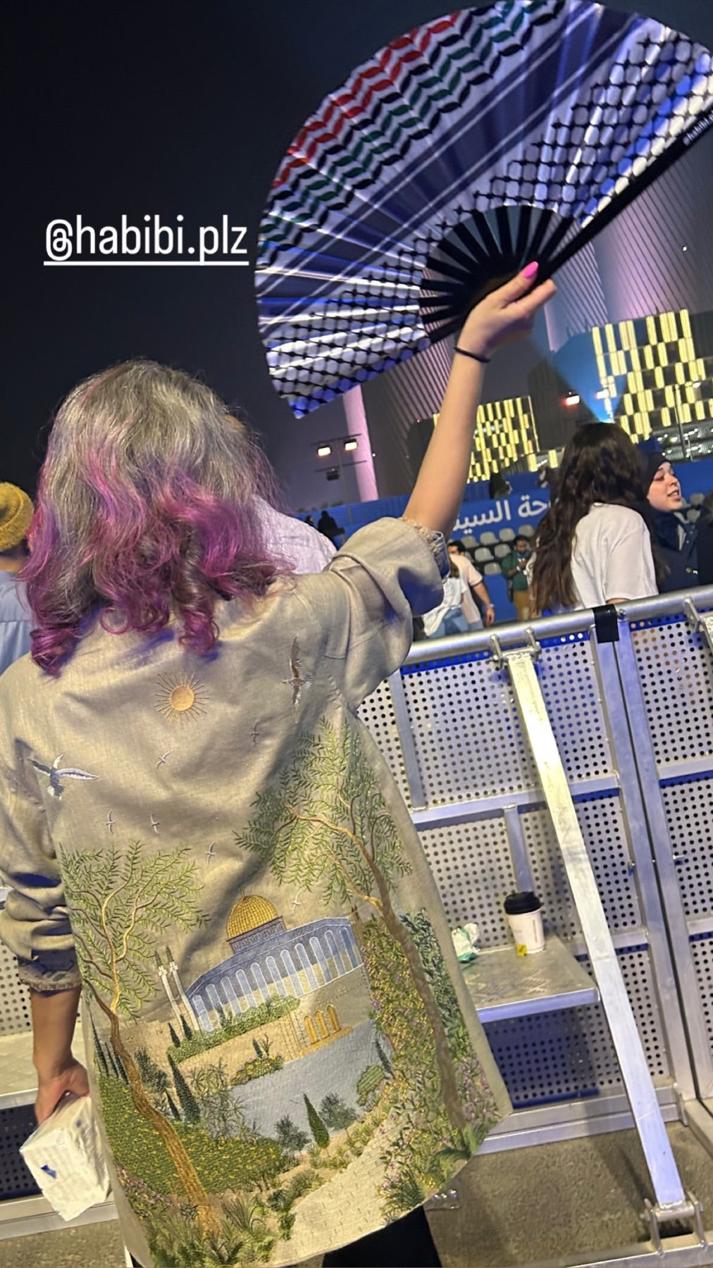 A woman raising a keffiyeh-pattern Habibi fan at a live event