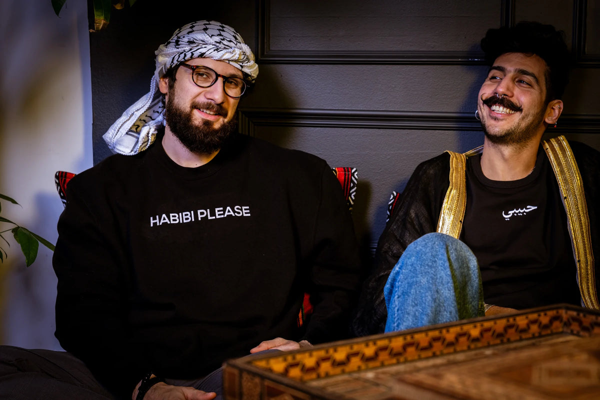 Two people sitting together, one wearing a HABIBI PLEASE sweatshirt